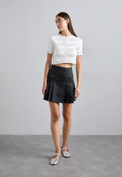 Jeune femme portant des ballerines argentées, un cardigan court à manches courtes blanc avec des détails en perles, et une mini-jupe plissée noire taille haute.