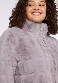 Jasnofioletowa kurtka z futerka ekologicznego z wysokim kołnierzem, zapięciem na zamek błyskawiczny oraz teksturowanym wzorem, charakteryzująca się miękką, pluszową powierzchnią.