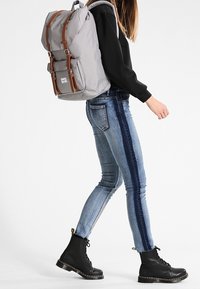 Sac à dos en toile grise avec des accents en cuir brun, des bretelles ajustables et une poche avant. Porté avec un jean bleu et des bottes noires à lacets.