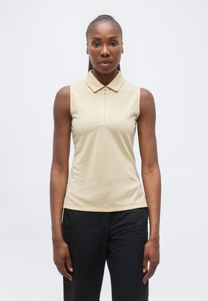 Žena s tmavou pokožkou a zviazanými vlasmi, ktorá má na sebe béžový bezrukávový top s golierom a predným zipsom a čierne nohavice na jednofarebnom bielom pozadí.