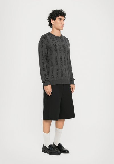 Grauer gestrickter Pullover mit Logo-Design, gerippten Bündchen und Saum. Trägt schwarze Shorts, weiße Socken und schwarze Slipper in stehender Pose.