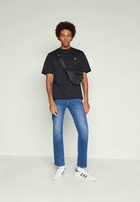 Young man with afro wearing black t-shirt, blue jeans, white sneakers, and black crossbody bag, standing against plain light background.