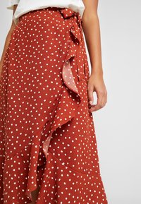 Gonna avvolgente di colore ruggine con pois bianchi, orlo arricciato e dettaglio di cintura annodata, realizzata in tessuto leggero.