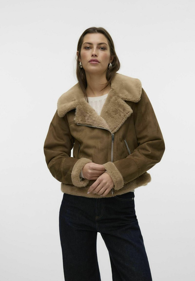 Giacca zip-up in suede marrone con colletto e polsini in pelliccia, caratterizzata da un design aderente e una texture morbida, abbinata a jeans scuri.