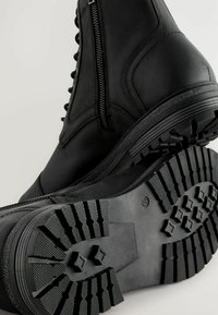 Botas de cuero negro con caña alta, que presentan un cierre lateral con cremallera y una suela de goma texturizada con un patrón de agarre para mayor tracción.