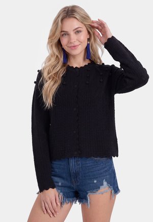 Lyshåret kvinde med langt bølget hår iført en sort tekstureret knaptrøje, blå frynseøreringe og slidte denimshorts, smiler blidt.