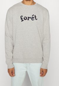 Lichtgrijze sweatshirt met zwarte tekst "forét" gedragen door een persoon in lichtblauwe jeans, met handen ontspannen aan de zijkanten, tegen een effen achtergrond.