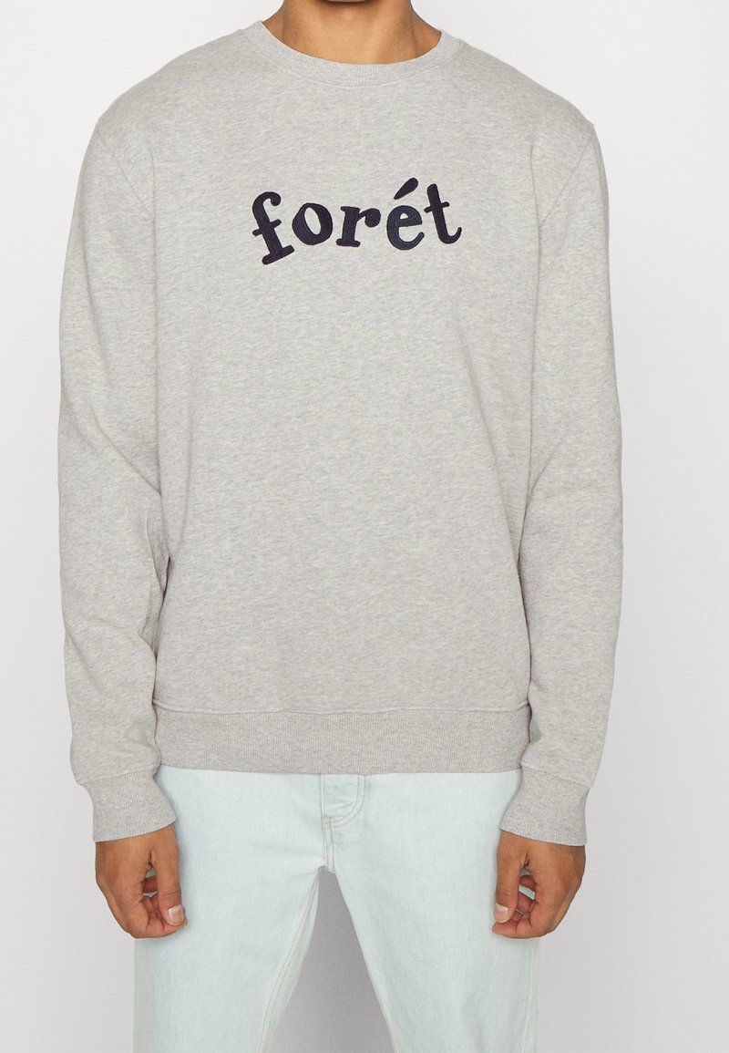 Lichtgrijze sweatshirt met zwarte tekst "forét" gedragen door een persoon in lichtblauwe jeans, met handen ontspannen aan de zijkanten, tegen een effen achtergrond.