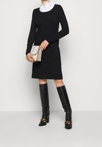 Robe noire à manches longues avec col rond, associée à des bottes en cuir noir hautes jusqu'aux genoux avec un accent en chaîne dorée et une pochette beige.