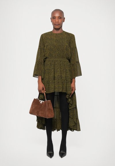 Mulher vestindo um vestido verde estampado em corte assimétrico, meia-calça preta, sapatos de bico fino pretos, segurando uma bolsa de camurça marrom com fecho prateado.