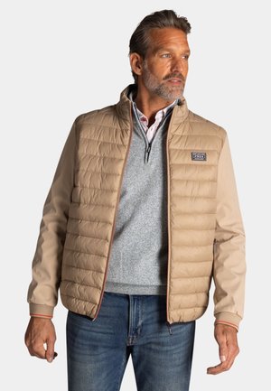 Man draagt een beige doorgestikte jas over een grijze trui met rits en blauwe jeans, kijkt opzij tegen een effen achtergrond.