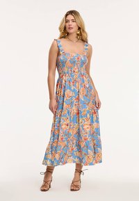 Ziedu maxi kleita gaiši zilā krāsā ar raupjām krūtīm, volāniem rotātiem plecu siksniņām, spilgtām oranžām un dzeltenām rakstu detaļām un plīvojošu A-līnijas siluetu.