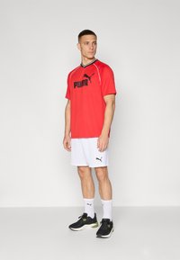 Rød Puma idrettsdrakt med svart logo og hvite detaljer, sammen med hvite shorts med en liten svart Puma-logo. Svart joggesko og hvite sokker.