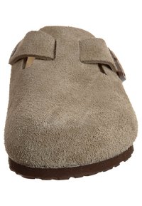 Scarpa slip-on in suede beige con due cinturini regolabili, punta rotonda e suola in gomma marrone scuro. Presenta una texture morbida e un design minimalista.