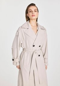 Jasno beżowy trencz z podwójnym zapięciem, dużym kołnierzem i bocznymi kieszeniami. Posiada pasek i mankiety zapinane na guziki.