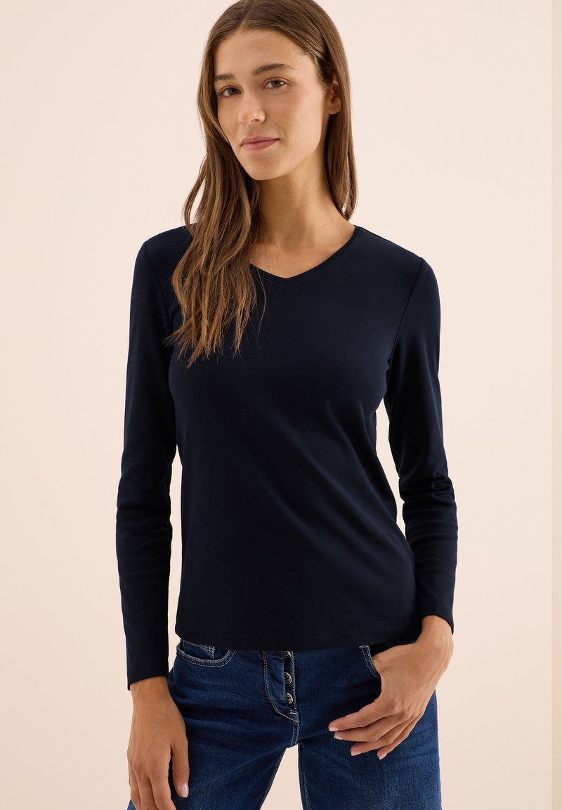 Marinefarbener Langarm-V-Ausschnittshirt aus weichem Stoff; figurbetonte Form mit glatter Textur. Getragen mit blauen Jeans mit Knopfakzenten.