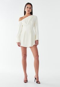 Abito mini bianco a spalla unica con maniche lunghe, corpetto strutturato e dettagli plissé. Tessuto morbido con una leggera lucentezza. Sandali con tacco alto.