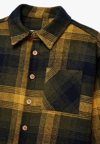 Rutet flannel skjorte i mørkegrøn, gul og marineblå; har spidse kraver, knaplukning foran og en brystlomme med et diagonalt mønster.