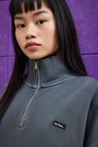 Ung kvinne med rett svart hår som har på seg en grå zip-up genser med «lets frans...» felt foran en lilla vegg.