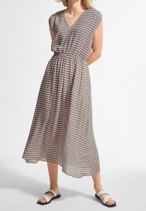 Femme portant une robe midi sans manches à motif, cintrée à la taille, et des sandales blanches à bouts ouverts, debout devant un fond uni.