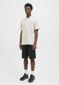 Beige polo shirt with short sleeves and collar, featuring a small logo. Paired with black cargo shorts and black sneakers.