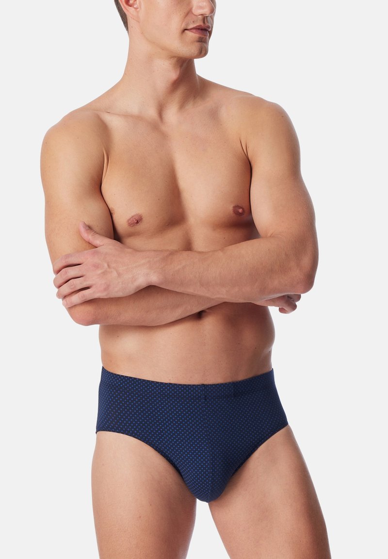 Oberkörperfreier Mann mit verschränkten Armen, der dunkelblaue Unterhosen mit einem subtilen Muster trägt, steht vor einem schlichten, hellen Hintergrund.