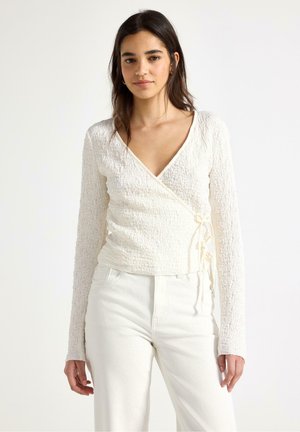 Ung kvinde iført tekstureret langærmet hvid top i omslagsstil og hvide bukser med høj talje, stående mod en ensfarvet baggrund.