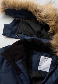 Mørkeblå isoleret jakke med en faux fur hættekant. Indersiden er sort og har en label med plejeinstruktioner og størrelsesdetaljer.