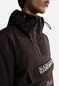 Brown anorak jacket with a high collar, front zip, and adjustable drawstrings. Features embroidered branding and a Norwegian flag patch.