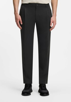 Homme portant un pantalon noir sur mesure et des baskets noires, debout devant un fond blanc uni.