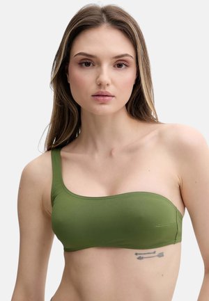 Giovane donna con lunghi capelli castani indossa un crop top verde monospalla, mostrando un tatuaggio a freccia sul lato destro sotto le costole.