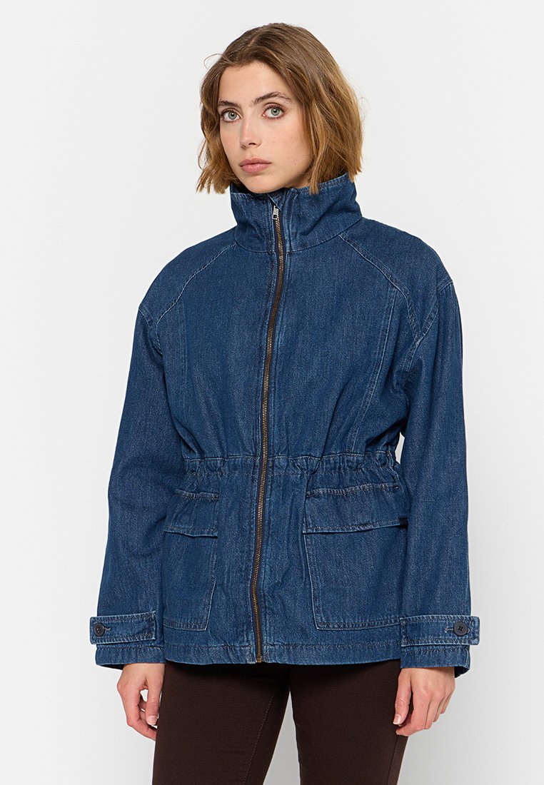 Chaqueta de mezclilla en azul oscuro, con un alto cuello, cremallera frontal, cintura ajustada y dos bolsillos frontales con tapetas.