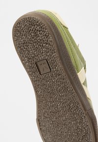 Η σόλα ενός πράσινου και μπεζ αθλητικού παπουτσιού που εμφανίζει υφασμένο καφέ λαστιχένιο πέλμα με ανάγλυφο τετράγωνο λογότυπο στο κέντρο.