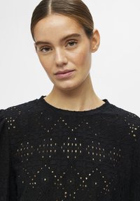 Haut en dentelle noire avec des motifs en diamants intriqués, un col rond et un tissu texturé transparent. Les manches bouffantes rehaussent les détails de conception.