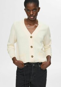 Cardigan color crema con escote en V, tres botones, puños acanalados y dos bolsillos frontales, hecho de un material de punto suave.