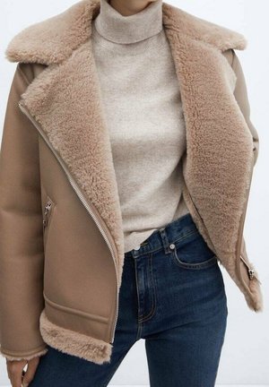 Frau trägt eine beige Lammfelljacke über einem beigen Rollkragenpullover und hoch taillierte dunkelblaue Jeans vor schlichtem Hintergrund.
