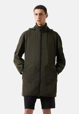 Man wearing olive green hooded long jacket with front zipper and black shorts, standing against plain light background.