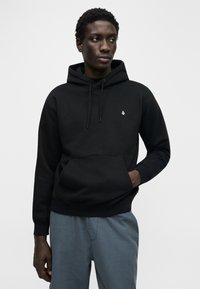 Μαύρο φούτερ με κουκούλα κατασκευασμένο από μαλακό ύφασμα, με μπροστινή τσέπη, ρυθμιζόμενα κορδόνια και μικρό λευκό λογότυπο στο στήθος.