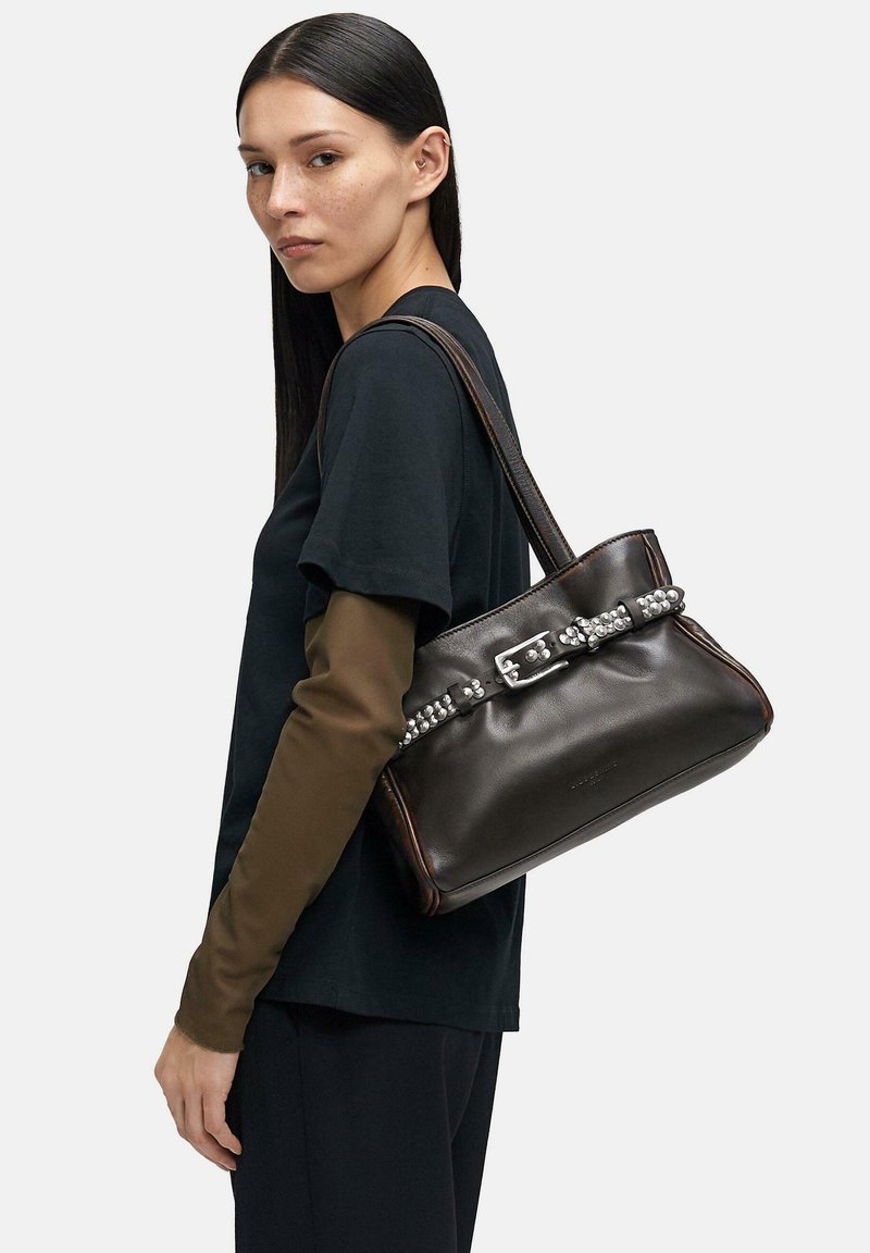 Czarna skórzana torebka z detalem w postaci srebrnej sprzączki, o strukturalnym kształcie, z brązowymi akcentami, trzymana przez osobę ubraną w czarną koszulkę i brązowe długie rękawy.