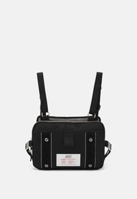 Musta kangaskassi säädettävillä hihnoilla, jossa on hopeanvärisiä yksityiskohtia, verkkokankaita, rohkeaa valkoista ommelta ja suuri brändilogo edessä.