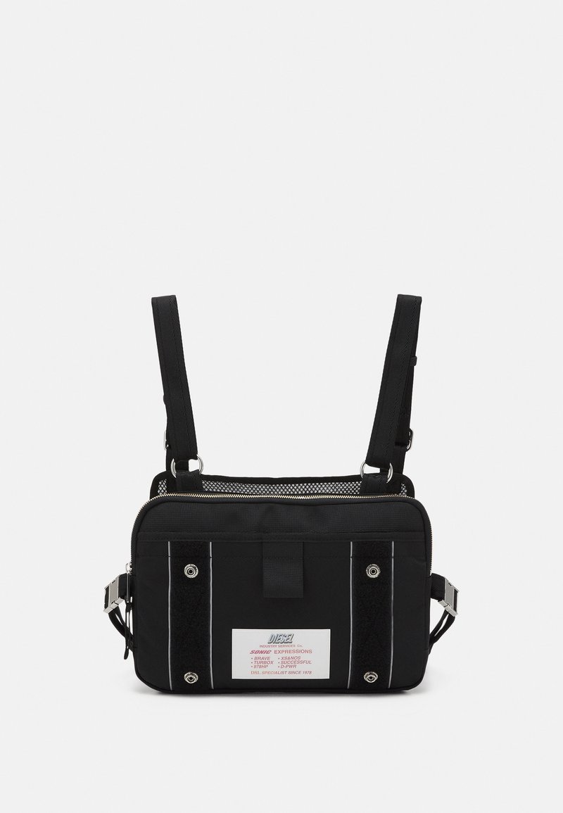 Musta kangaskassi säädettävillä hihnoilla, jossa on hopeanvärisiä yksityiskohtia, verkkokankaita, rohkeaa valkoista ommelta ja suuri brändilogo edessä.