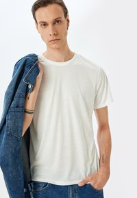 Weißes T-Shirt mit Rundhalsausschnitt aus weichem Stoff, kombiniert mit einer blauen Jeansjacke. Die Arme des Models sind sichtbar, mit einem Tattoo am linken Handgelenk.