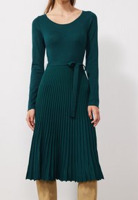 Abito a pieghe verde acqua con maniche lunghe, scollatura rotonda e cintura in vita. La parte superiore liscia si trasforma in pieghe texturizzate nella gonna.