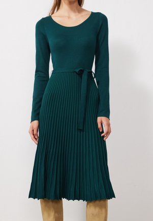 Robe plissée bleu sarcelle avec manches longues, col rond et ceinture à la taille. La partie supérieure lisse se transforme en plis texturés au niveau de la jupe.