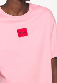 Różowa bawełniana koszulka z okrągłym dekoltem. Posiada prostokątną, czerwoną naszywkę z haftem, na której widnieje napis „HUGO” w kolorze czarnym. Krótkie rękawy.