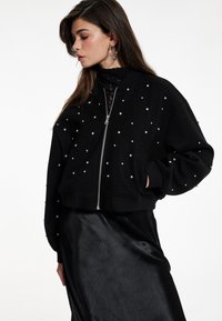 Schwarze Bomberjacke mit Reißverschluss vorne, verziert mit kleinen, funkelnden Nieten. Sie hat einen gerippten Saum und Bündchen und wird mit einem eleganten schwarzen Rock kombiniert.