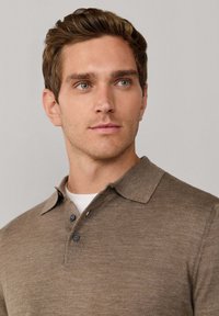 Aansluitend bruin poloshirt van zachte stof, met een klassieke kraag, drieknoopsluiting en subtiele textuur. Neutrale onderlaag zichtbaar.