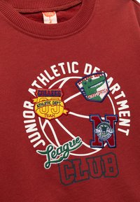 Sudadera burdeos con cuello redondo que presenta parches bordados en blanco y colores, incluyendo un diseño de baloncesto y varios logotipos de departamentos deportivos.