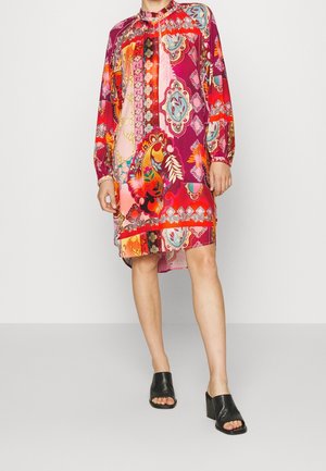 Kvinde iført en knælængde kjole med lange ærmer, dekoreret med livlige blomster- og geometriske mønstre i røde, pink, orange og blå nuancer, kombineret med sorte åbne tå-sandaler.