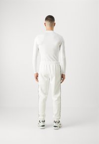 Bílá upnutá košile s dlouhými rukávy v kombinaci s odpovídajícími bílými tepláky s černými bočními pruhy a černobílými sportovními botami.